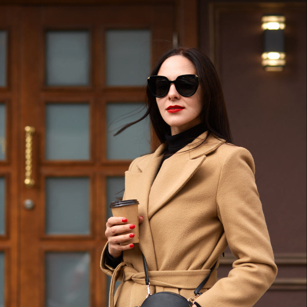 Woman in a beige coat holding a coffee cup in front of a wooden door.